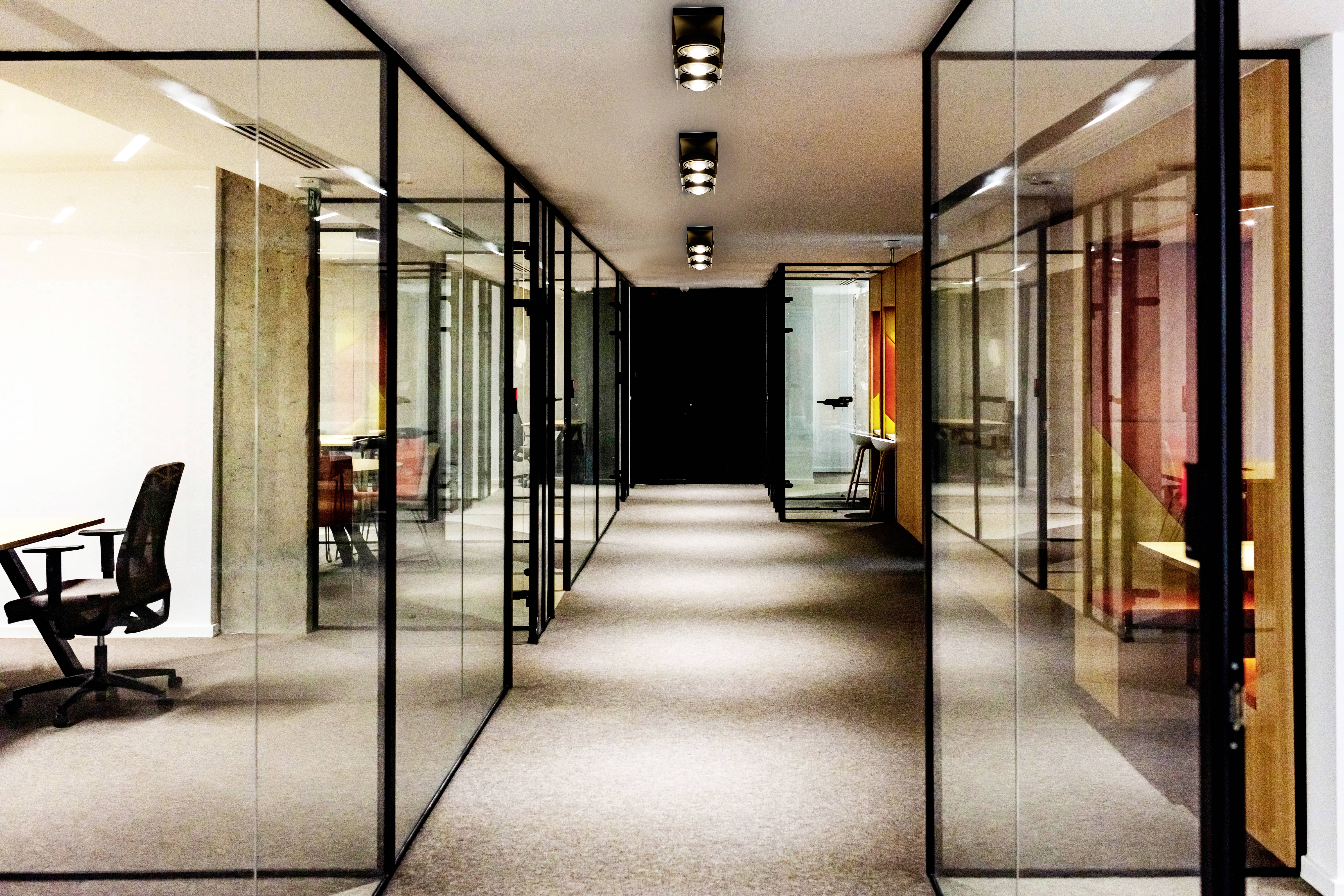 Długi korytarz nowoczesnego biura z przeszklonymi drzwiami i ściankami, białymi lampami sufitowymi oraz pojedynczymi pustymi pomieszczeniami biurowymi.