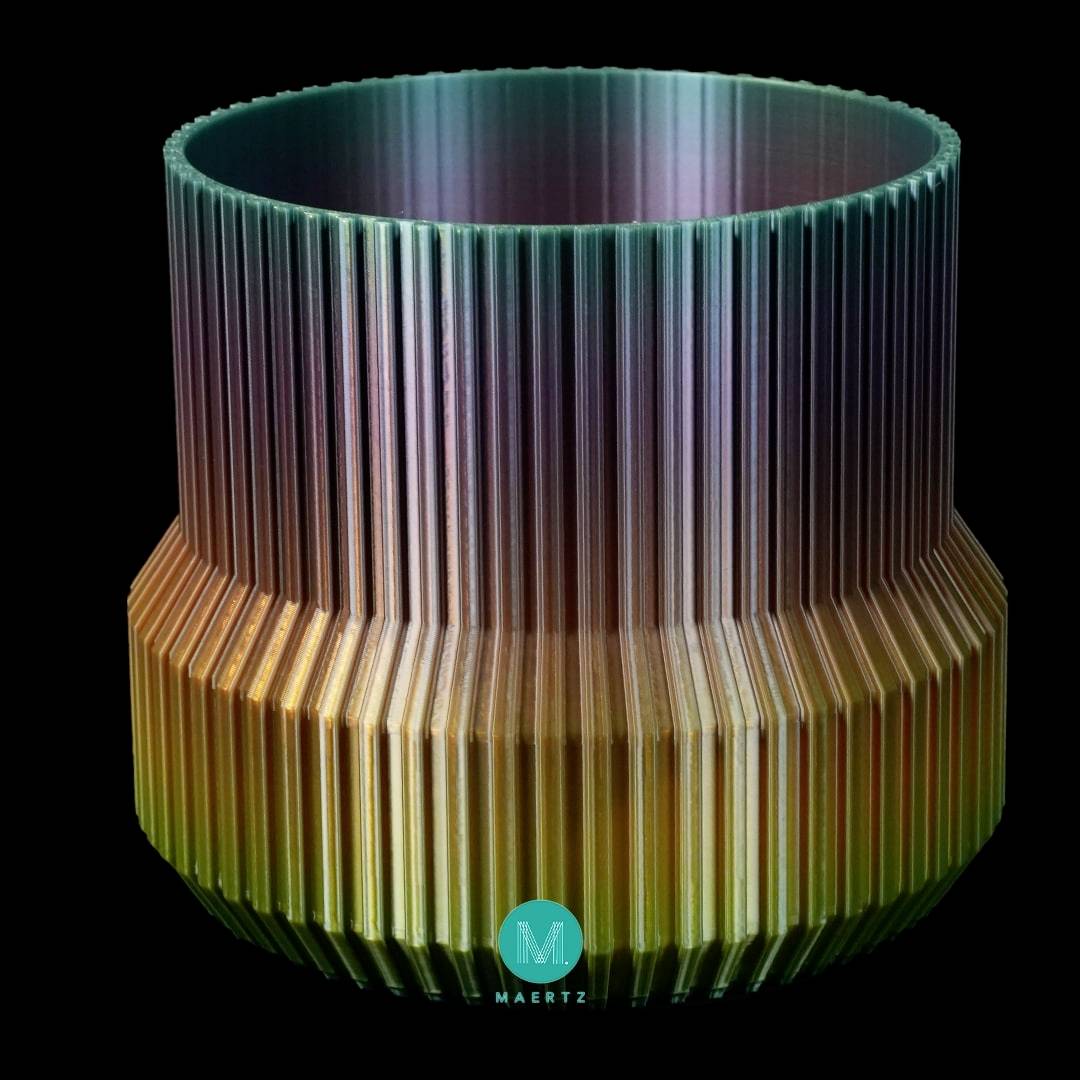 Cylindryczny obiekt z falistymi, przechodzącymi w gradiencie kolorami rowkami, od zieleni na górze do pomarańczu na dole. Logo 'M' widoczne u dołu.