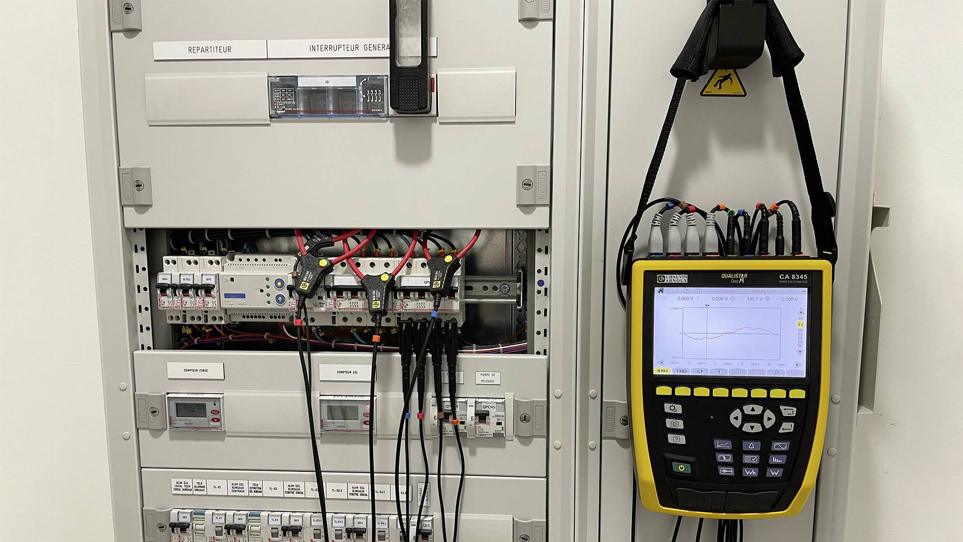 Elektryczny rozdzielnik elektryczny z otwartą przednią ścianką ukazuje przewodzone komponenty. Przyrząd pomiarowy wisi po prawej stronie i wyświetla wykres na ekranie.