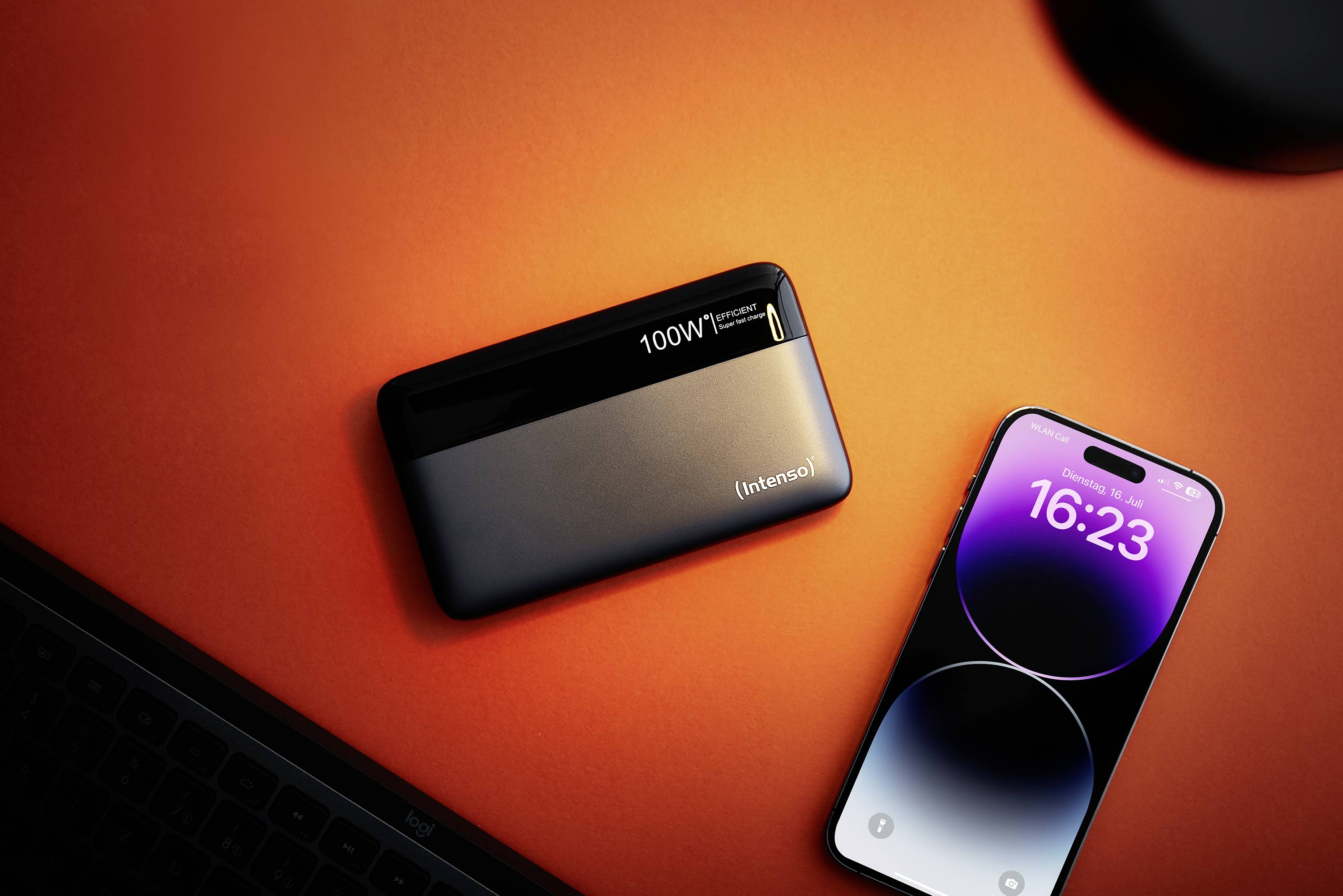 Smartfon leży obok czarnego powerbanku na pomarańczowym stole. Cyfrowy czas na smartfonie wskazuje 16:23.