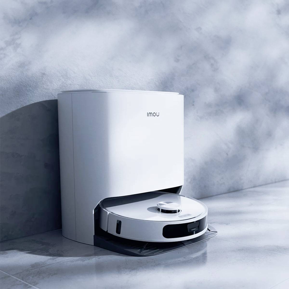 Biały robot-odkurzacz ze stacją ładowania przy ścianie, oświetlony światłem słonecznym padającym z lewej strony na podłogę.