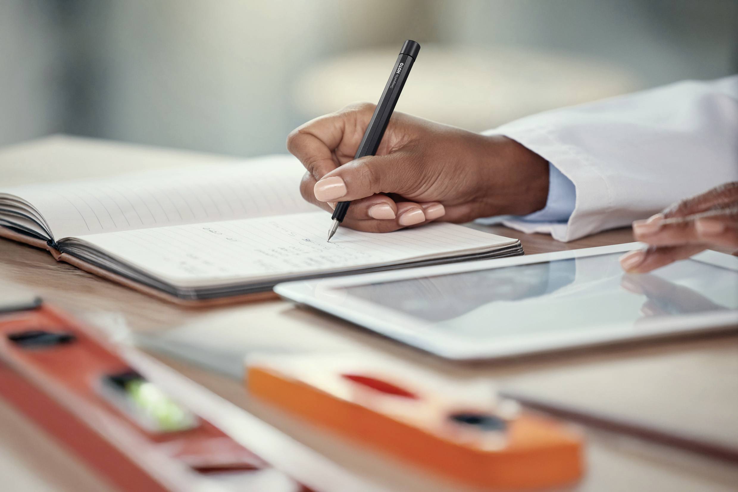 Osoba pisze notatki w zeszycie obok tabletu leżącego na stole. Na pierwszym planie widoczne są poziomnica i linijka.