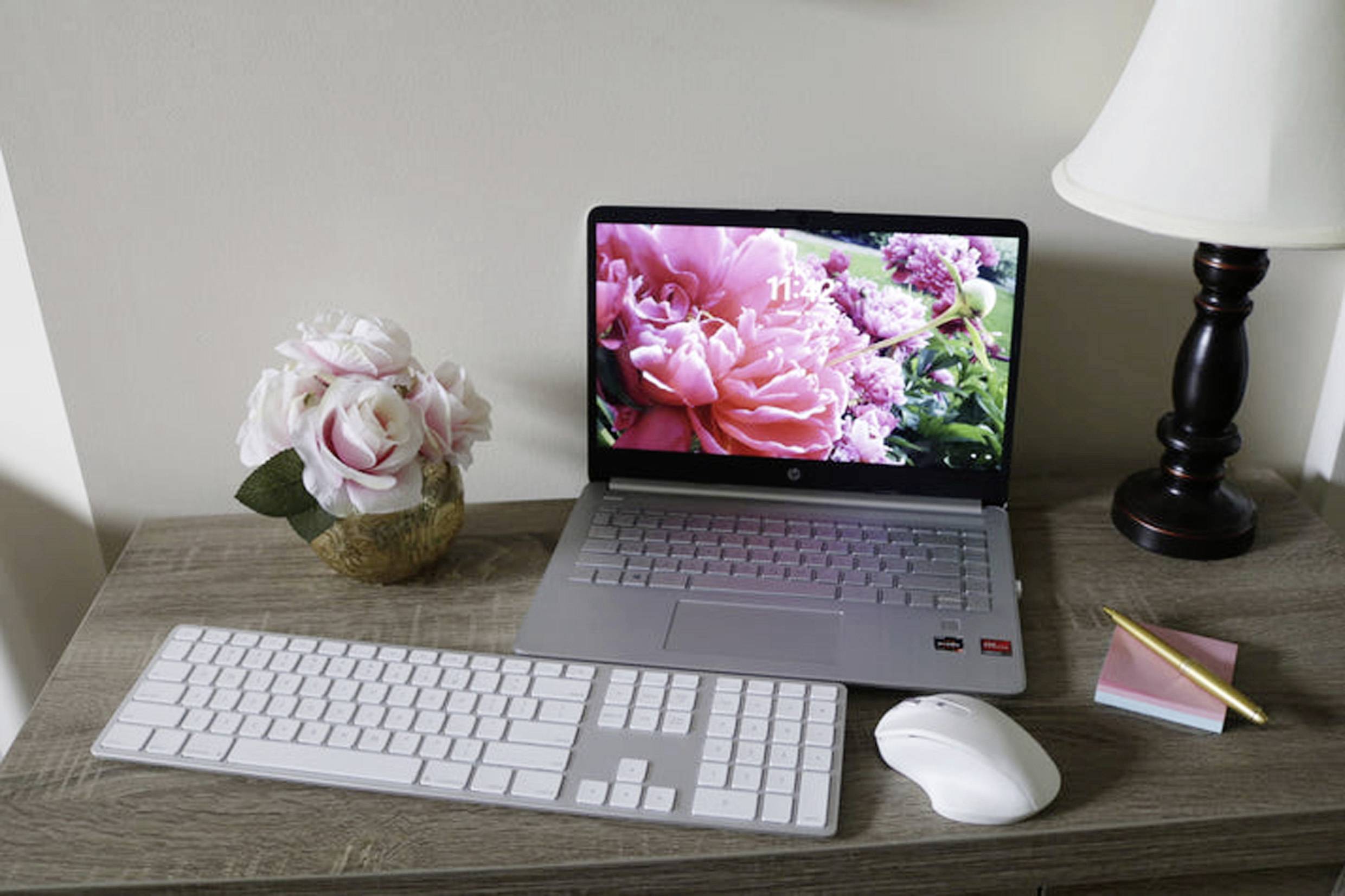 Laptop z ekranem w kwiatki stoi na biurku obok klawiatury, myszy, notatnika, ołówka, lampki biurkowej i wazonu z kwiatami.