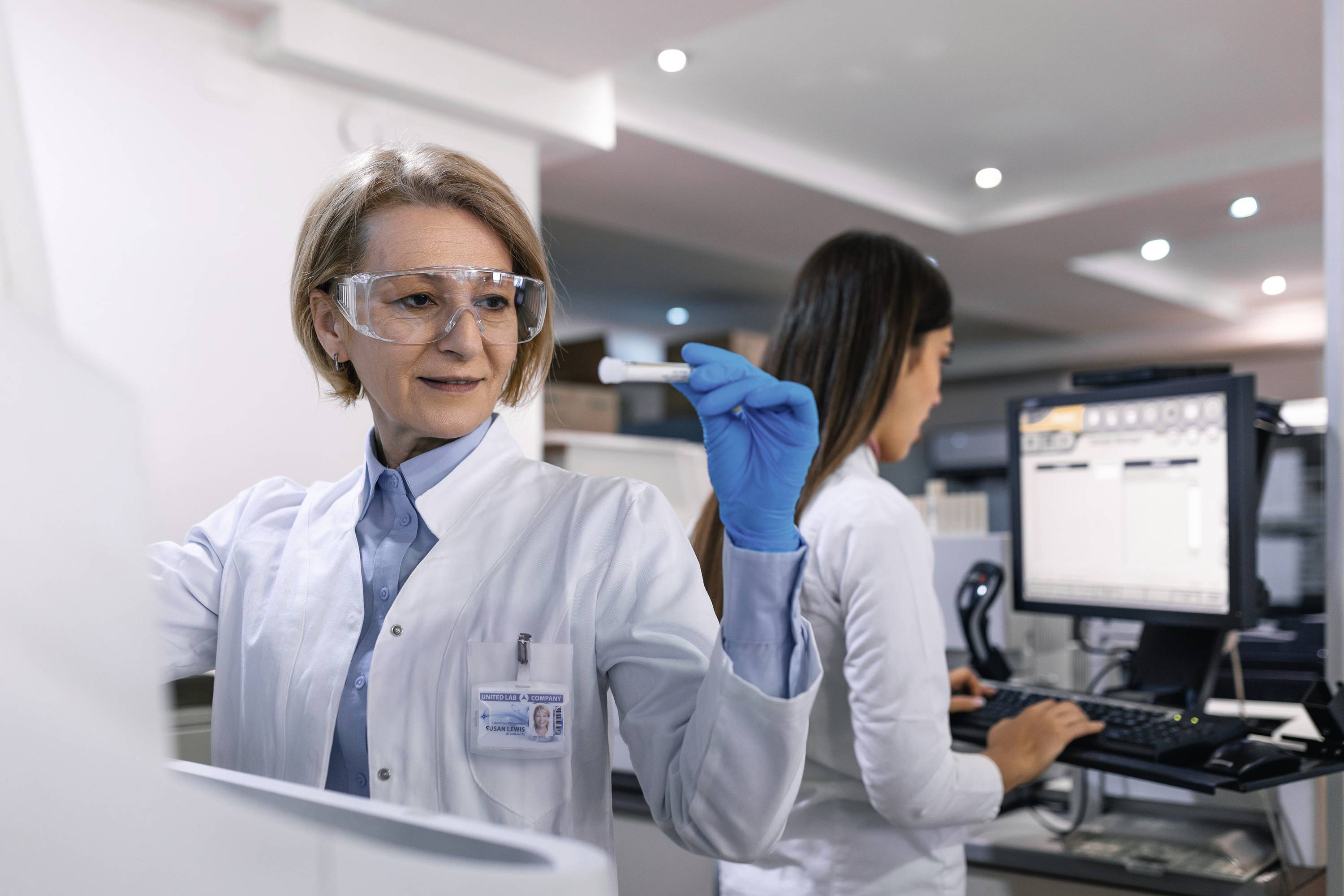 Dwie naukowczynie w laboratorium; ta z przodu nosi okulary ochronne i rękawiczki, trzyma pipetę. W tle: komputer z danymi.
