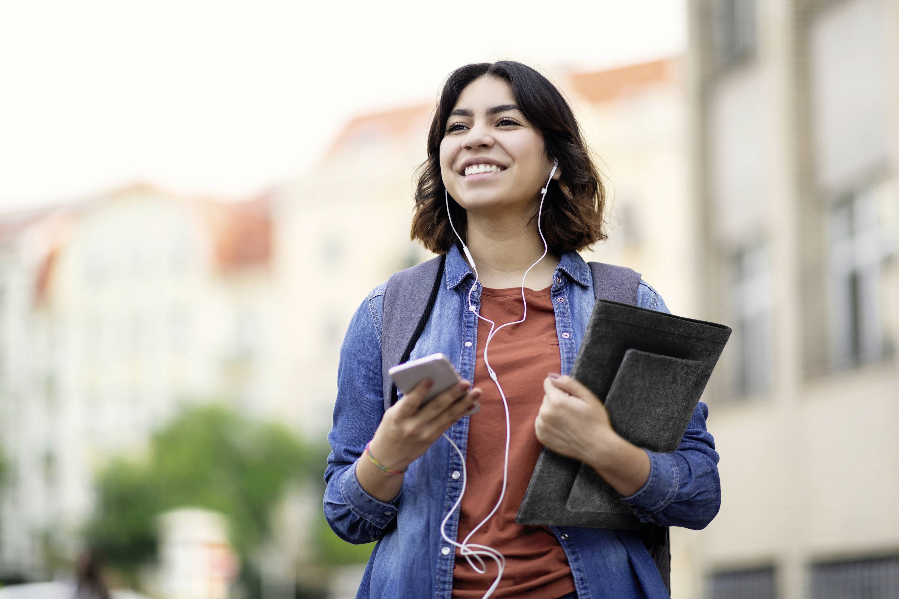 Kobieta uśmiecha się na dworze, ma słuchawki, trzyma smartfona i ciemnoszary notes.