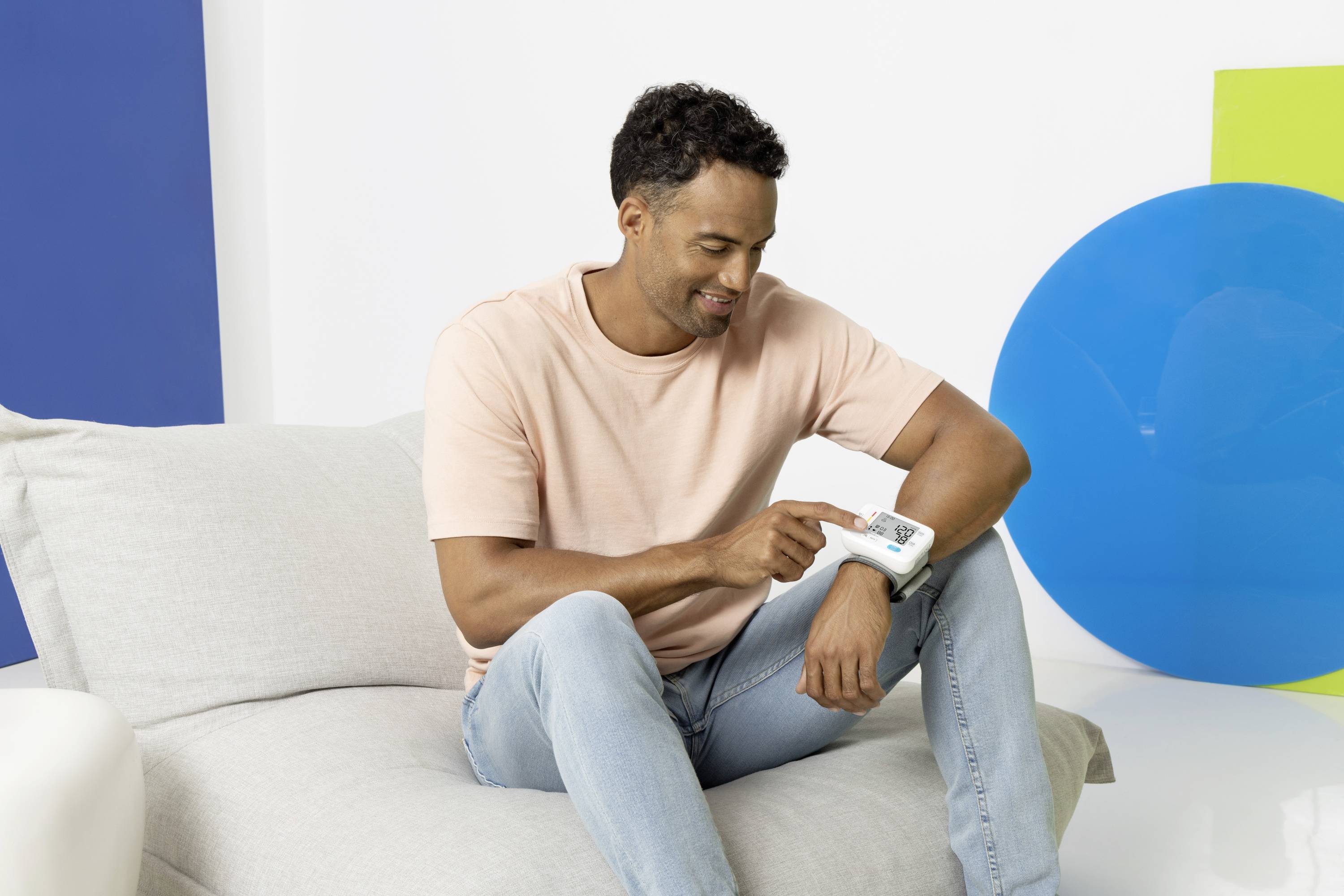 Mężczyzna w różowej koszulce i dżinsach siedzi na kanapie i przygląda się urządzeniu trzymanemu w ręku. Kolorowe kształty w tle.