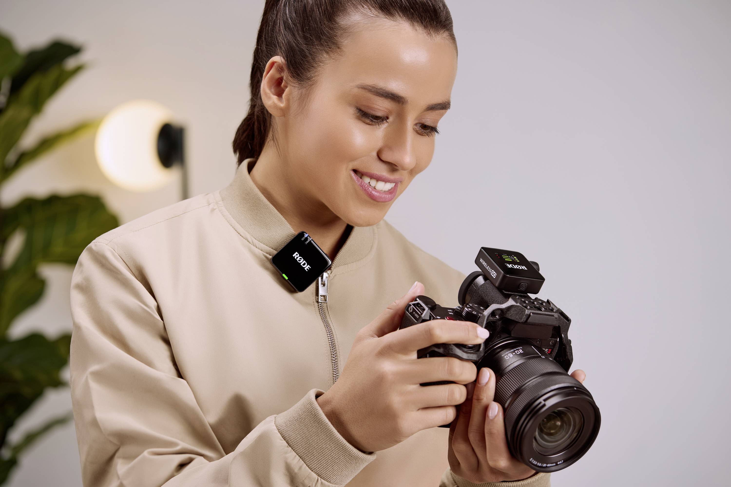 Osoba trzyma profesjonalny aparat fotograficzny i się uśmiecha. Ma na sobie piaskowy top i mikrofon. W tle widać rośliny.