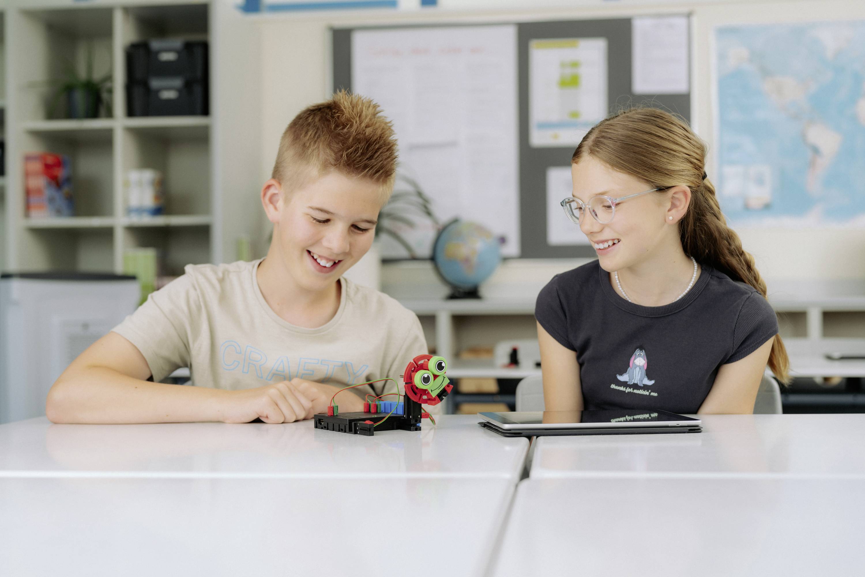 Chłopiec i dziewczynka siedzą przy stoliku w klasie, uśmiechają się i bawią małym robotem. Na tle widoczny jest globus.