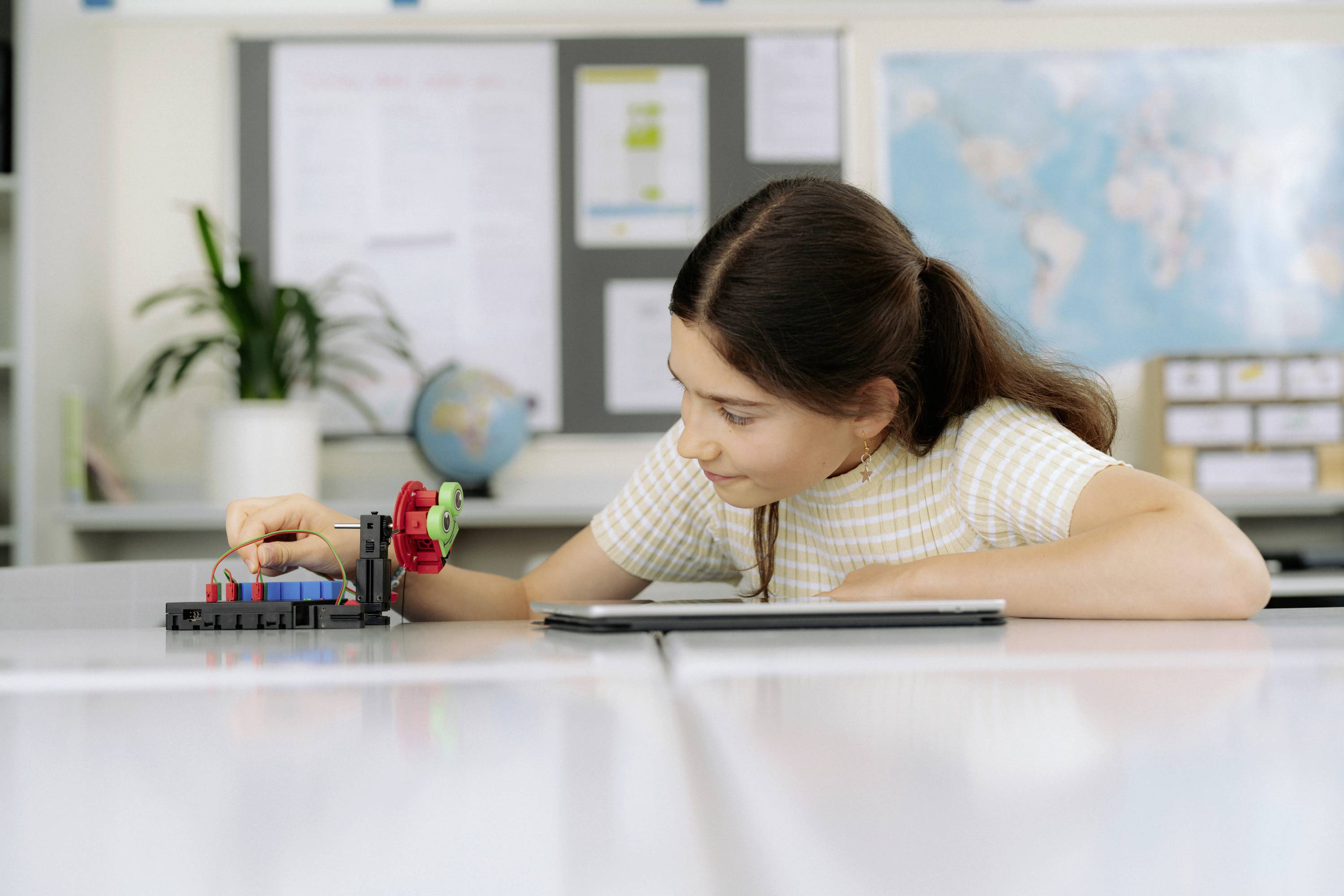 Dziewczyna koncentruje się na modelu technicznym na stole w klasie, w tle widać mapę świata i roślinę.
