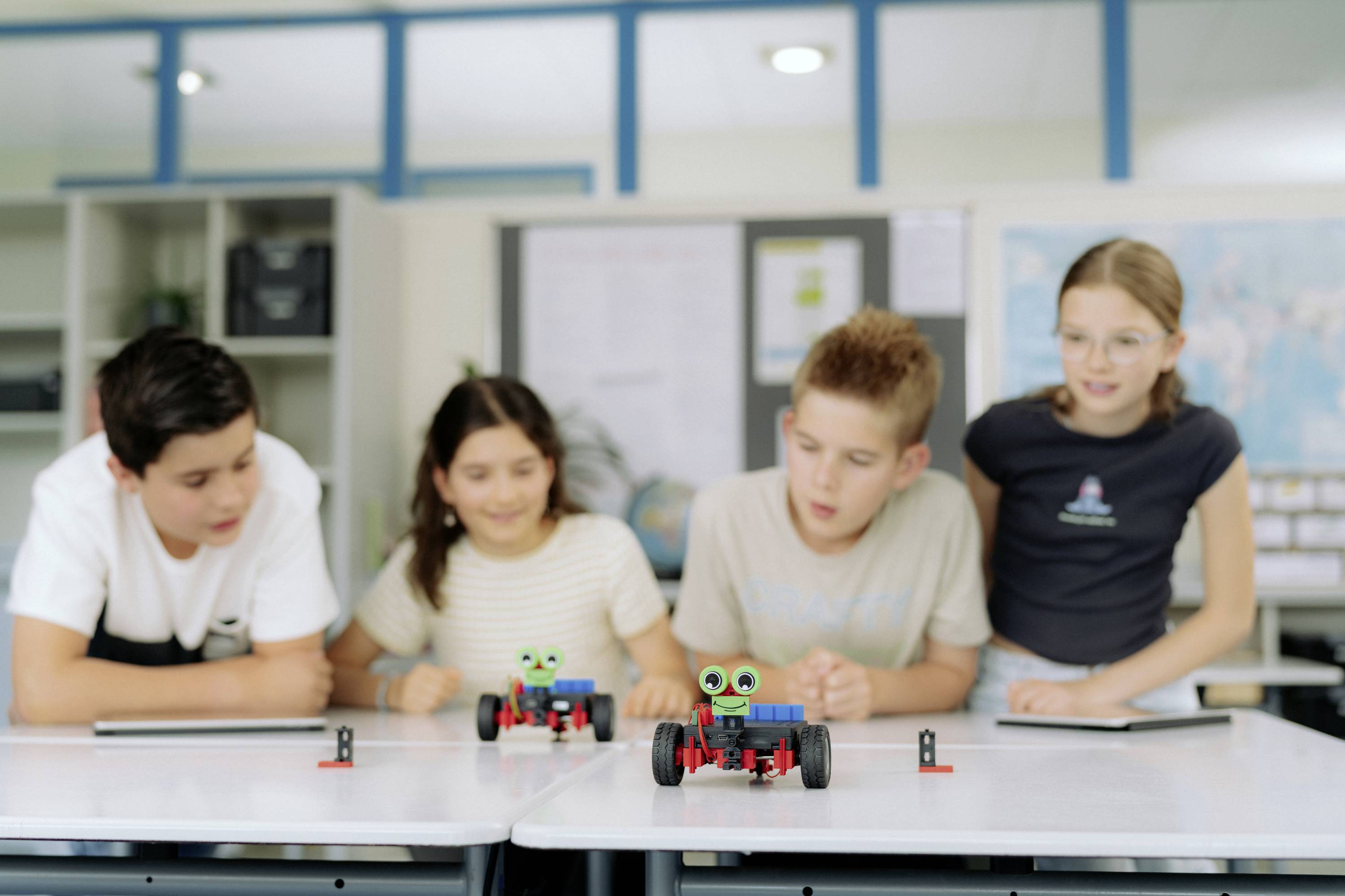 Czworo dzieci patrzy na dwa miniaturowe roboty stojące na stole w klasie. Wyglądają na skupionych i zainteresowanych. W tle widać półki i mapę świata.