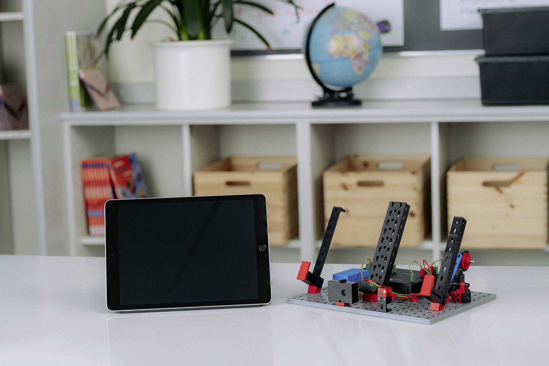 Tablet i mały model techniczny stoją na stole w sali lekcyjnej. W tle widać glob.