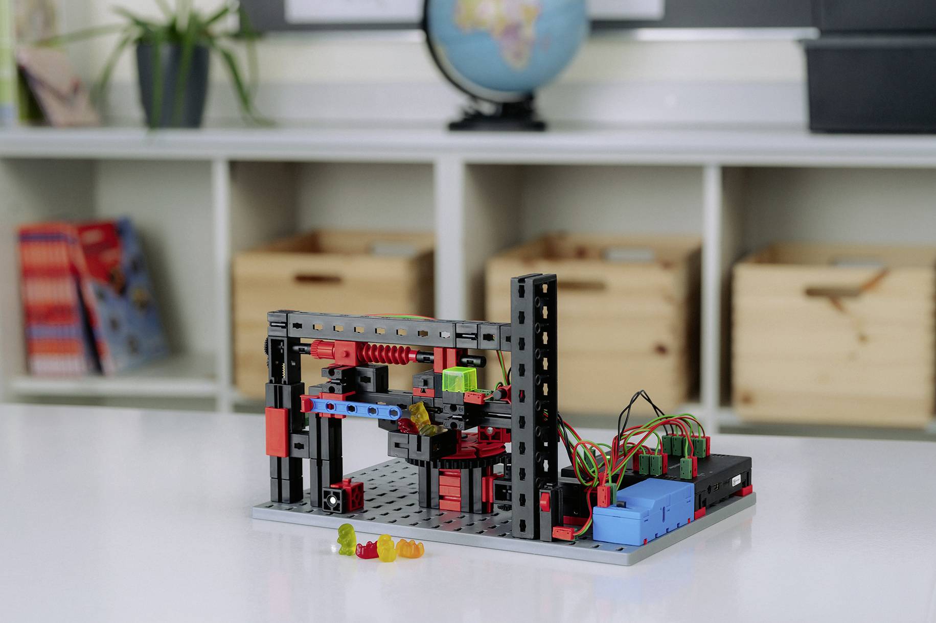 Robot Lego stojący na stole w sali lekcyjnej, otoczony kolorowymi żelkami. W tle widać regał z pudełkami.