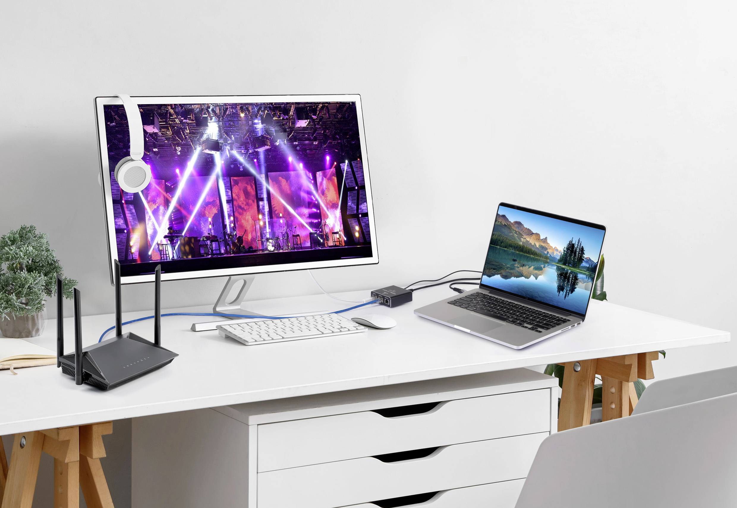 Nowoczesne biurko z monitorem wyświetlającym kolorowe przedstawienie sceniczne, laptopem z ekranem przedstawiającym przyrodę, klawiaturą i routerem.