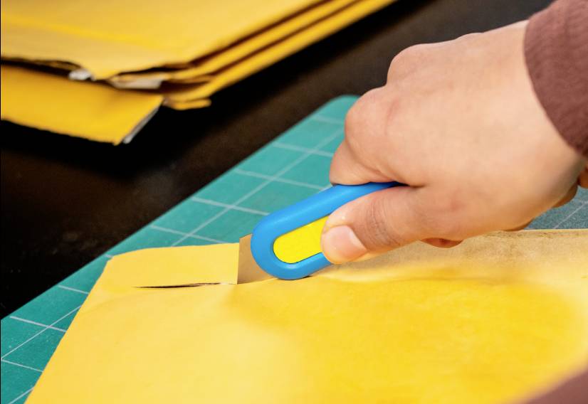 Jedna ręka trzyma niebieski nóż do tapet i nacina brązową kopertę na zielonej desce do krojenia.