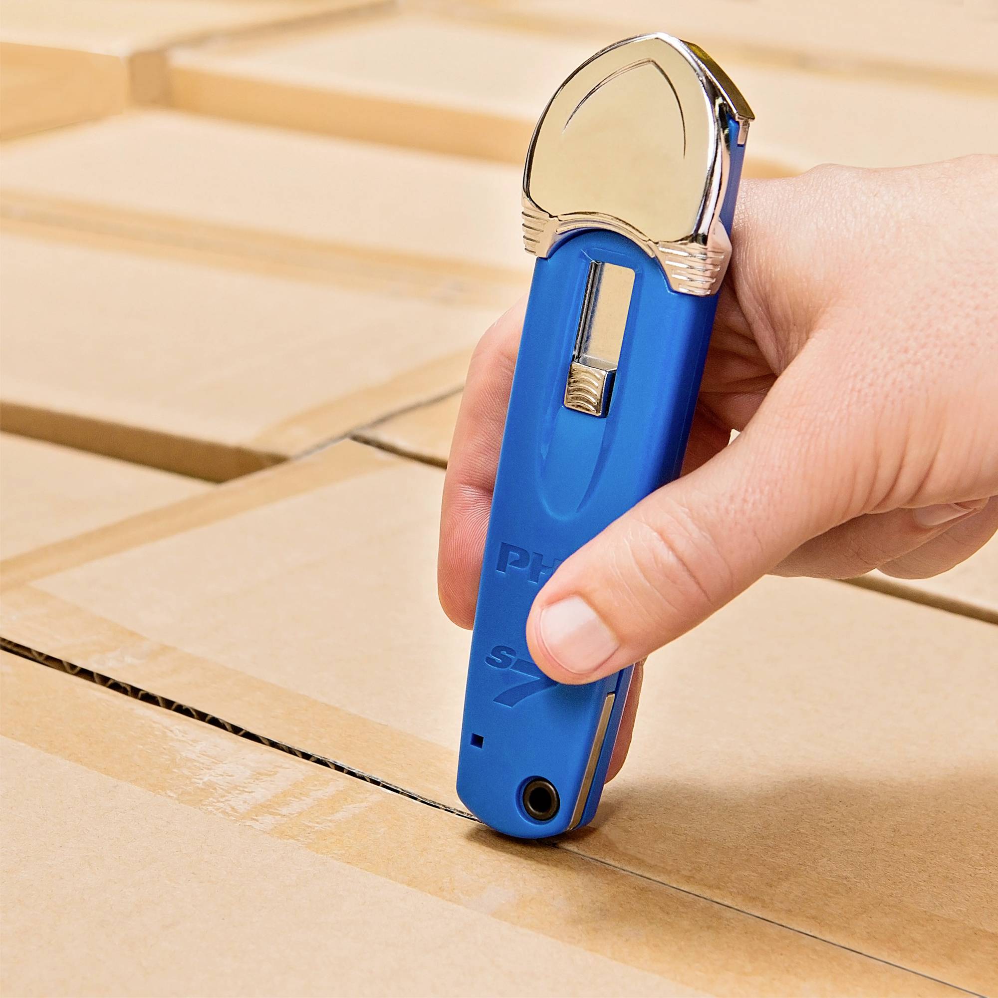 Dłoń trzyma niebieski nóż do tektury nad pudełkiem kartonowym, gotowa do cięcia. Karton jest starannie złożony i zamknięty.