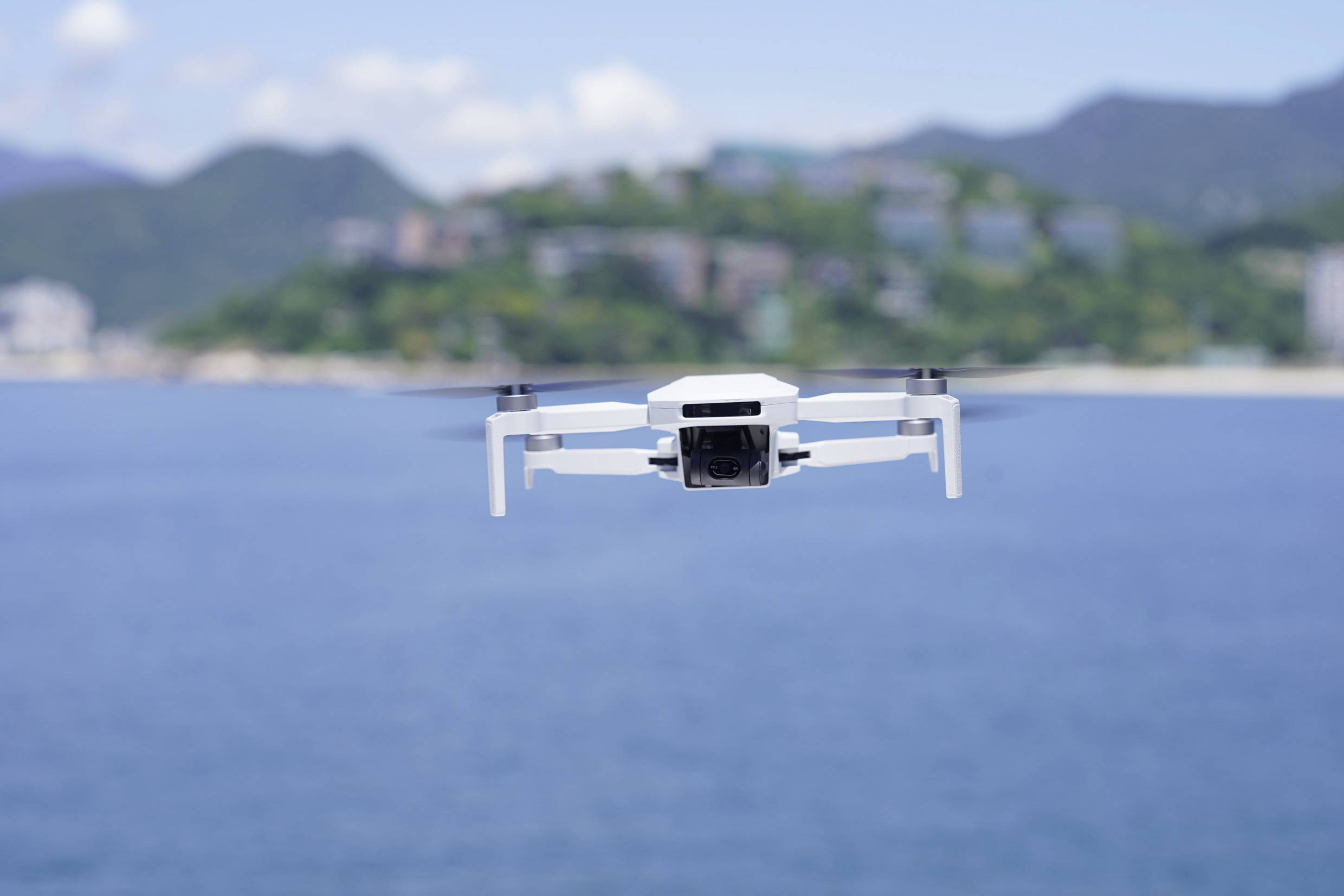 Biały dron unosi się nad niebieską wodą, a w tle niewyraźne zielone wzgórza.