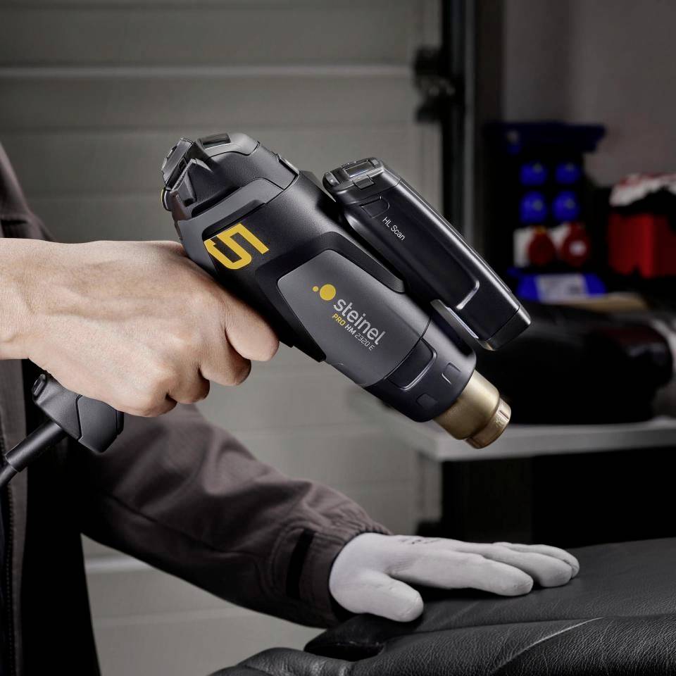 Osoba trzyma czarną pistolet na gorące powietrze marki Steinel, model HL 2020 E, w środowisku warsztatu.