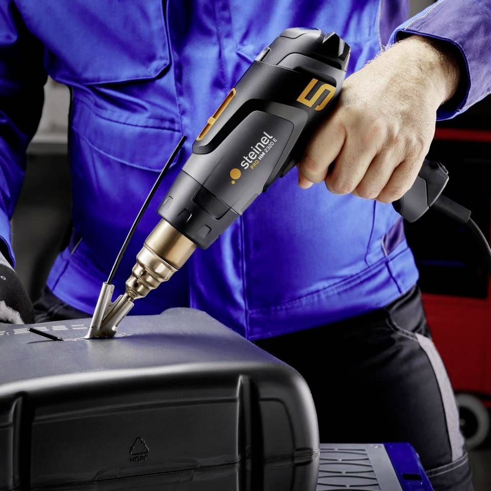 Pracownik używa pistoletu na gorące powietrze do obróbki elementu z tworzywa sztucznego. Ma na sobie niebieskie ubranie robocze.