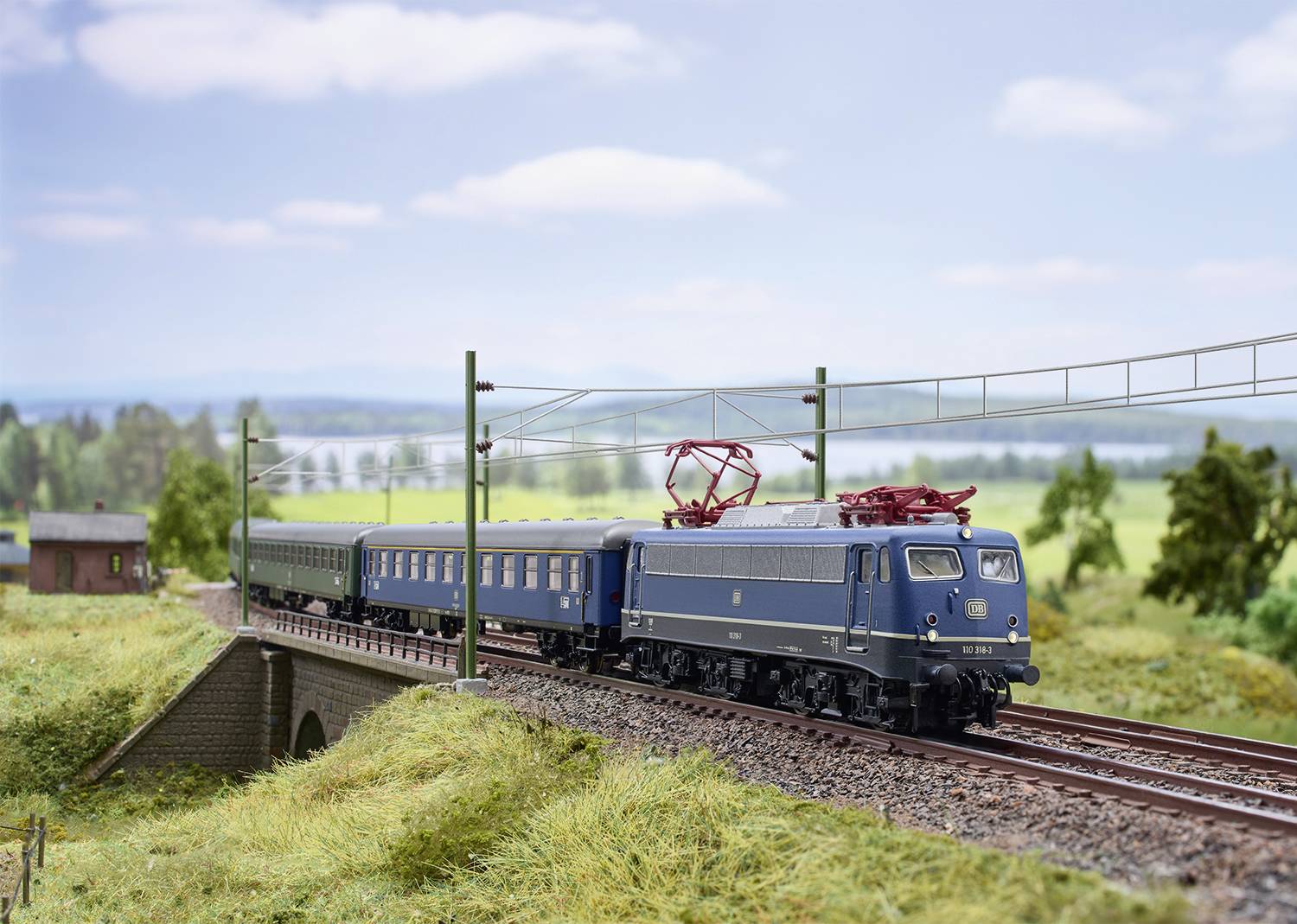 Niebieskoszary pociąg jedzie przez most w wiejskim krajobrazie. W tle widać pola i pochmurne niebo.