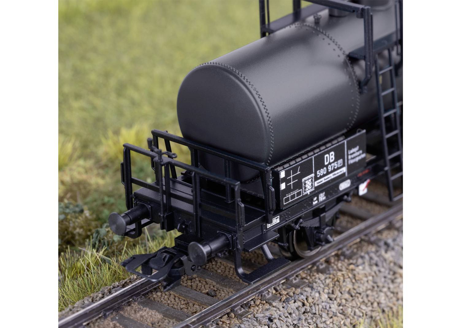 Model szarego wagonu towarowego Deutsche Bahn z precyzyjnie wykonanymi tabliczkami i szynami, otoczony trawą.