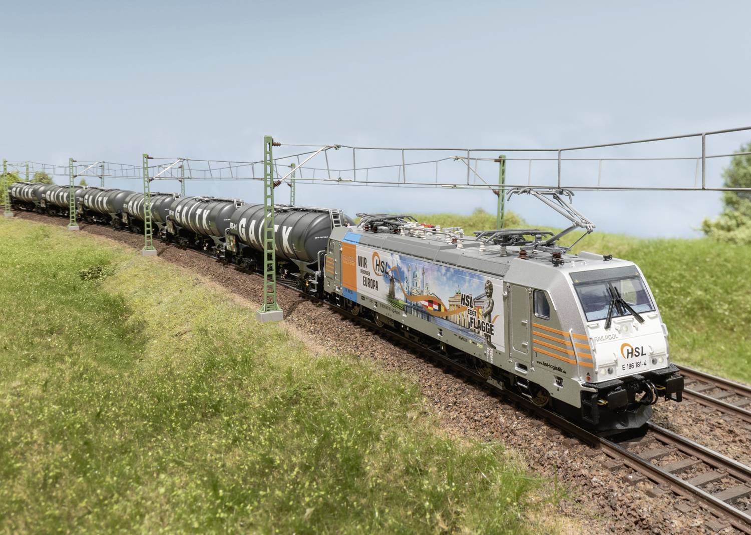 Pociąg towarowy jedzie po dwutorowym odcinku przez zieloną krajobraz. Skład składa się z kilku cystern.