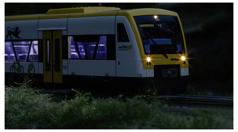 Mały modelowy pociąg elektryczny, żółty i biały, oświetlony, podróżujący po miniaturowych torach przez krajobraz pokryty trawą.