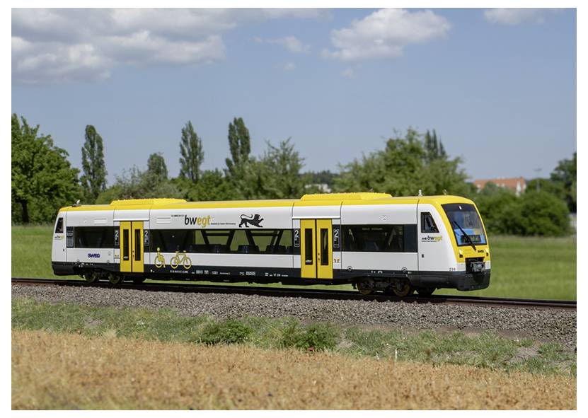 Żółto-biały pociąg podróżuje po torach przez trawiasty teren z drzewami i chmurami w tle.