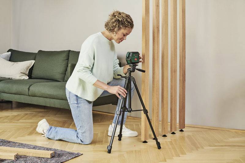 Osoba klęczy na podłodze i ustawia laser poziomujący na statywie w salonie, w pobliżu zielonej kanapy.
