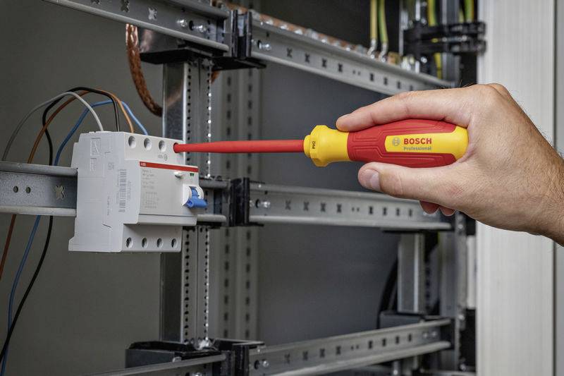 Dłoń dokręca wkręt izolowanym śrubokrętem marki Bosch na obudowie elektrycznego rozdzielnika.