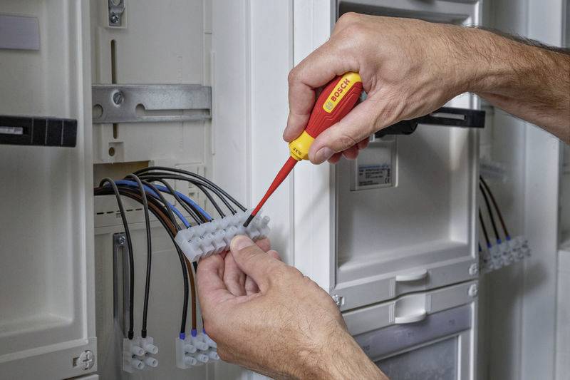 Mężczyzna używa śrubokręta do zamocowania przewodów w skrzynce bezpieczników. Nacisk kładziony jest na prawidłową i bezpieczną instalację.