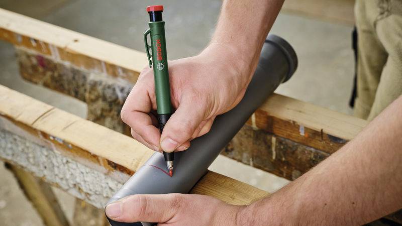 Dwie ręce zaznaczają zieloną kredką Bosch linię na szarym rurze leżącym na drewnianym podłożu.
