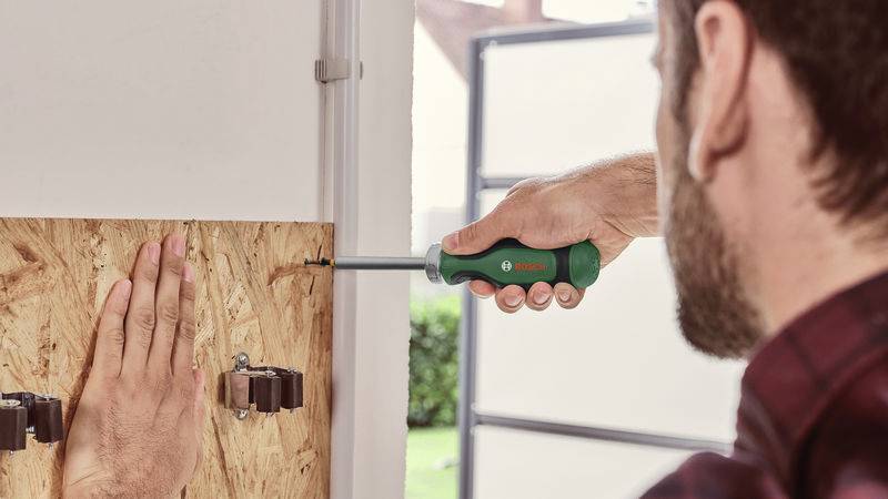 Osoba wkręca śrubę wkrętakiem w drewnianą płytę. Ręka podtrzymuje płytę, w tle widać okno.