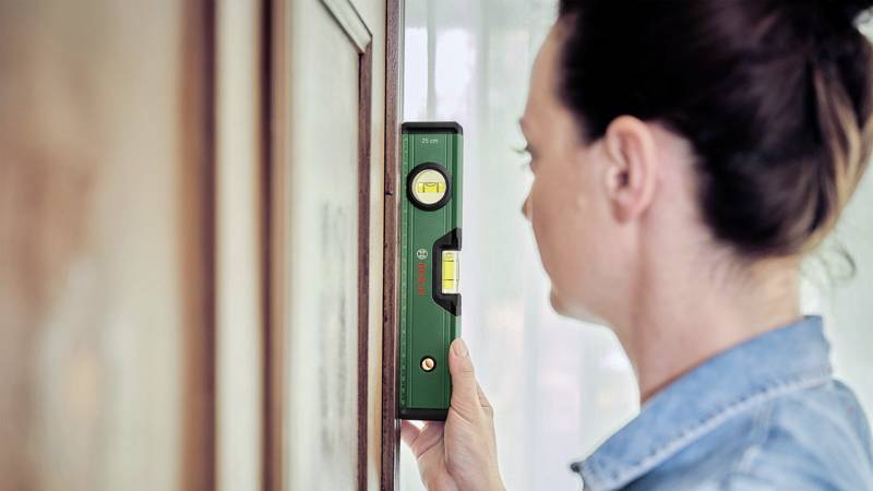 Kobieta sprawdza za pomocą zielonego poziomnika, czy drewniana deska jest ustawiona pionowo. Ma na sobie dżinsową koszulę.