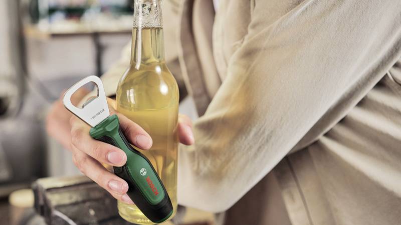 Osoba otwiera butelkę za pomocą elektrycznego otwieracza. Fokus jest skierowany na ruch otwierania.