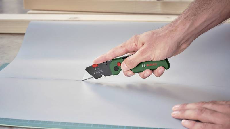 Ręka tnie szare materiał nożem do dywanów na macie do cięcia. Nóż posiada zielony uchwyt i czerwony przycisk.