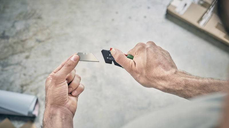 Osoba ostrzy żyletkę o metalową powierzchnię. Ręce są w centrum uwagi. W tle niewyraźne są pudła.