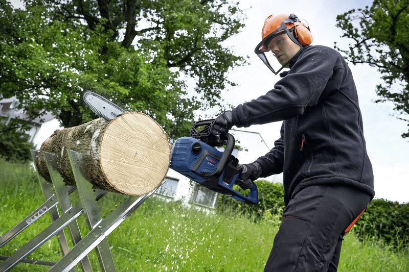 Osoba w odzieży ochronnej piłuje piłą mechaniczną pień drzewa na koźle tartacznym, wokół której rozciąga się zielona trawa i drzewa.