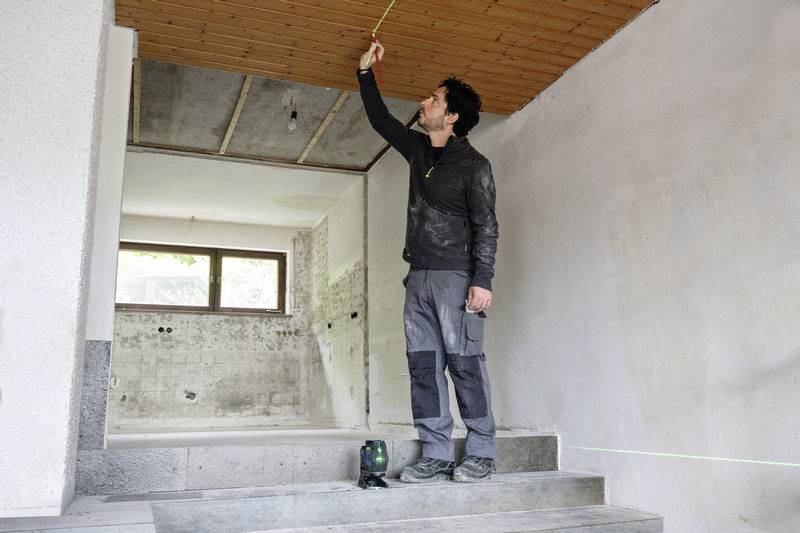 Mężczyzna mierzy wysokość sufitu w odnowionym pomieszczeniu za pomocą urządzenia laserowego. Ściany i sufit są jeszcze nieukończone.