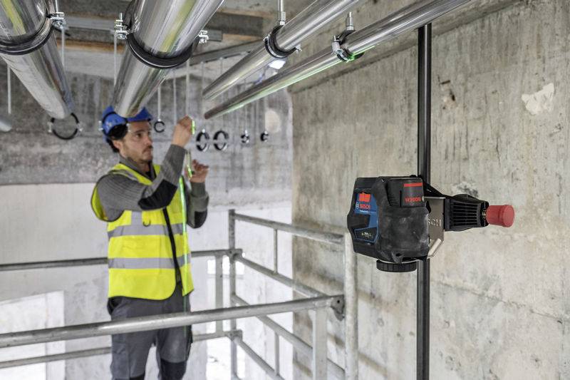 Mężczyzna w obowiązkowym wyposażeniu ochronnym pracuje na rusztowaniu przy metalowej rurze. Urządzenie laserowe rzutuje linię na rurociąg.