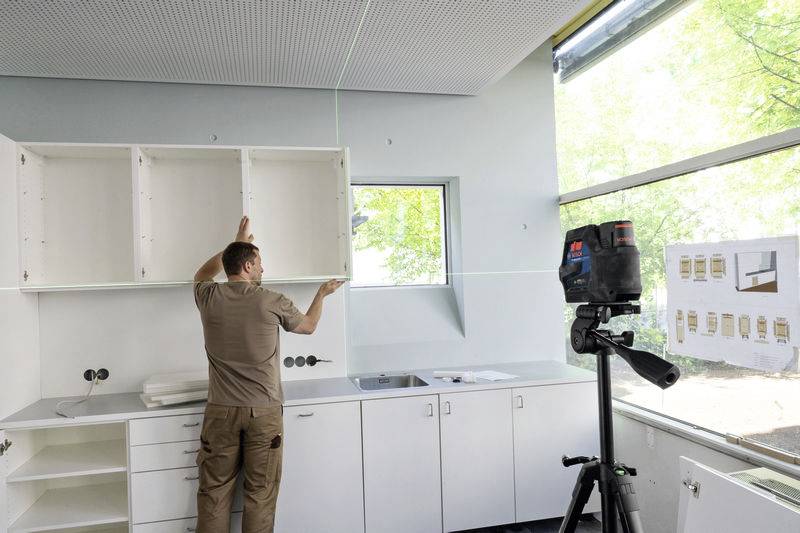 Mężczyzna montuje szafki kuchenne, podczas gdy urządzenie laserowe do poziomowania projektuje linię poziomą. W tle znajduje się duża przestrzeń okienna.