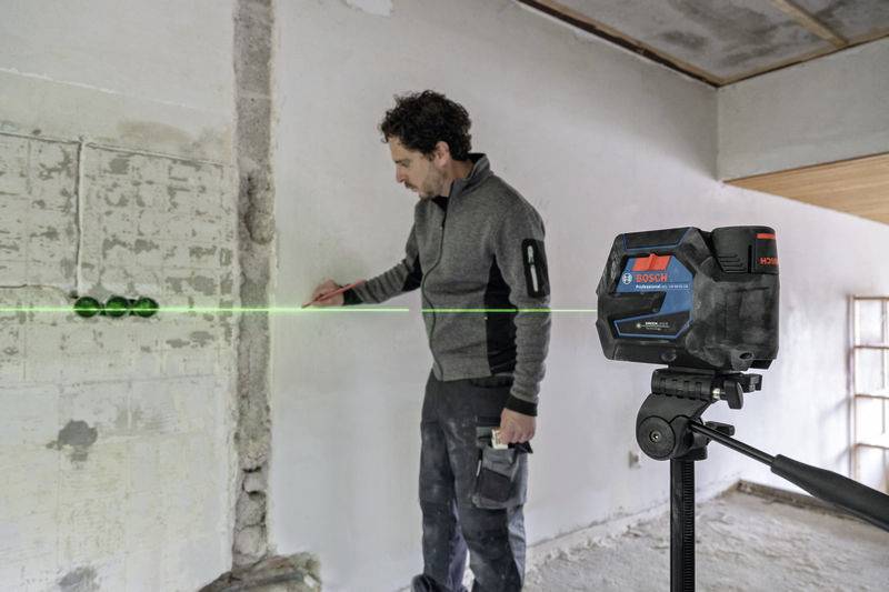 Mężczyzna używa laserowego urządzenia marki Bosch, aby oznaczyć prostą linię na ścianie w pomieszczeniu będącym w trakcie remontu.