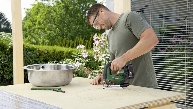 Mężczyzna wycina wyrzynarką okrągły otwór w desce. Metalowa miska stoi obok na stole.