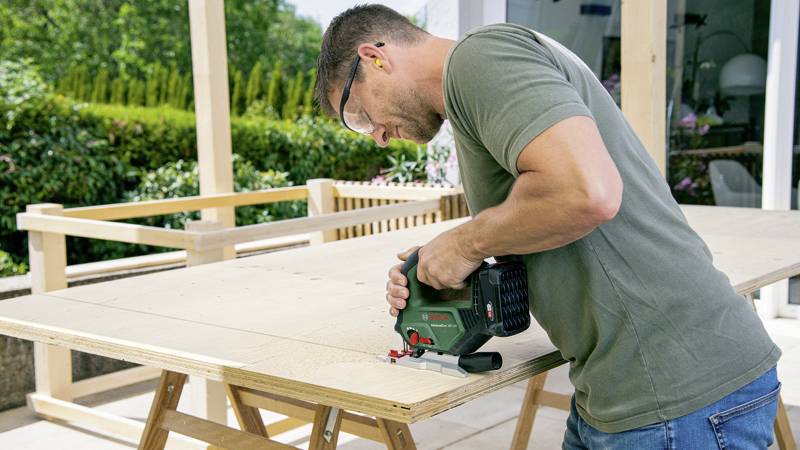 Mężczyzna w okularach ochronnych i nausznikach ochronnych przycina drewnianą płytę elektryczną wyrzynarką na tarasie.