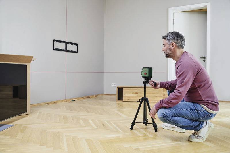 Mężczyzna reguluje laserowe urządzenie pomiarowe na statywie w salonie z drewnianą podłogą, aby precyzyjnie ustalić lokalizację telewizora.