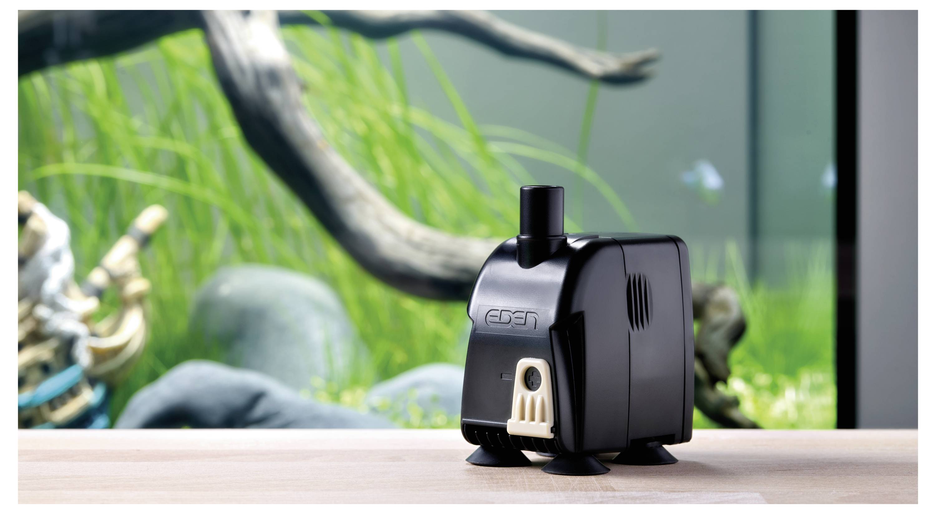 Mała czarna pompka akwariowa leży na drewnianej powierzchni przed tłem akwarium z zielonymi roślinami i drewnem niesiołowanym.