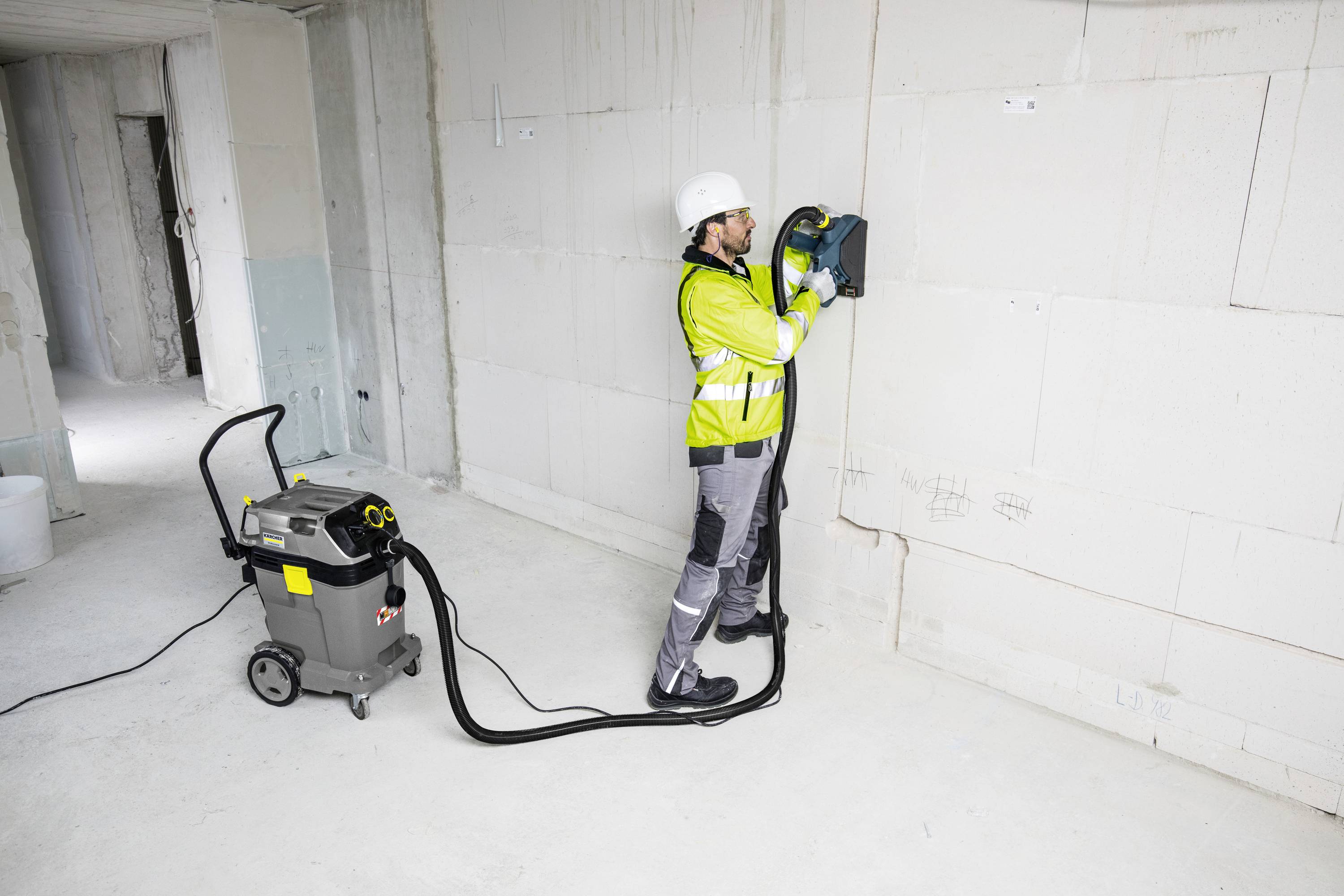 Robotnik budowlany w odzieży ochronnej obsługuje frezarkę do muru przy ścianie w niedokończonym pomieszczeniu. Odkurzacz jest podłączony.