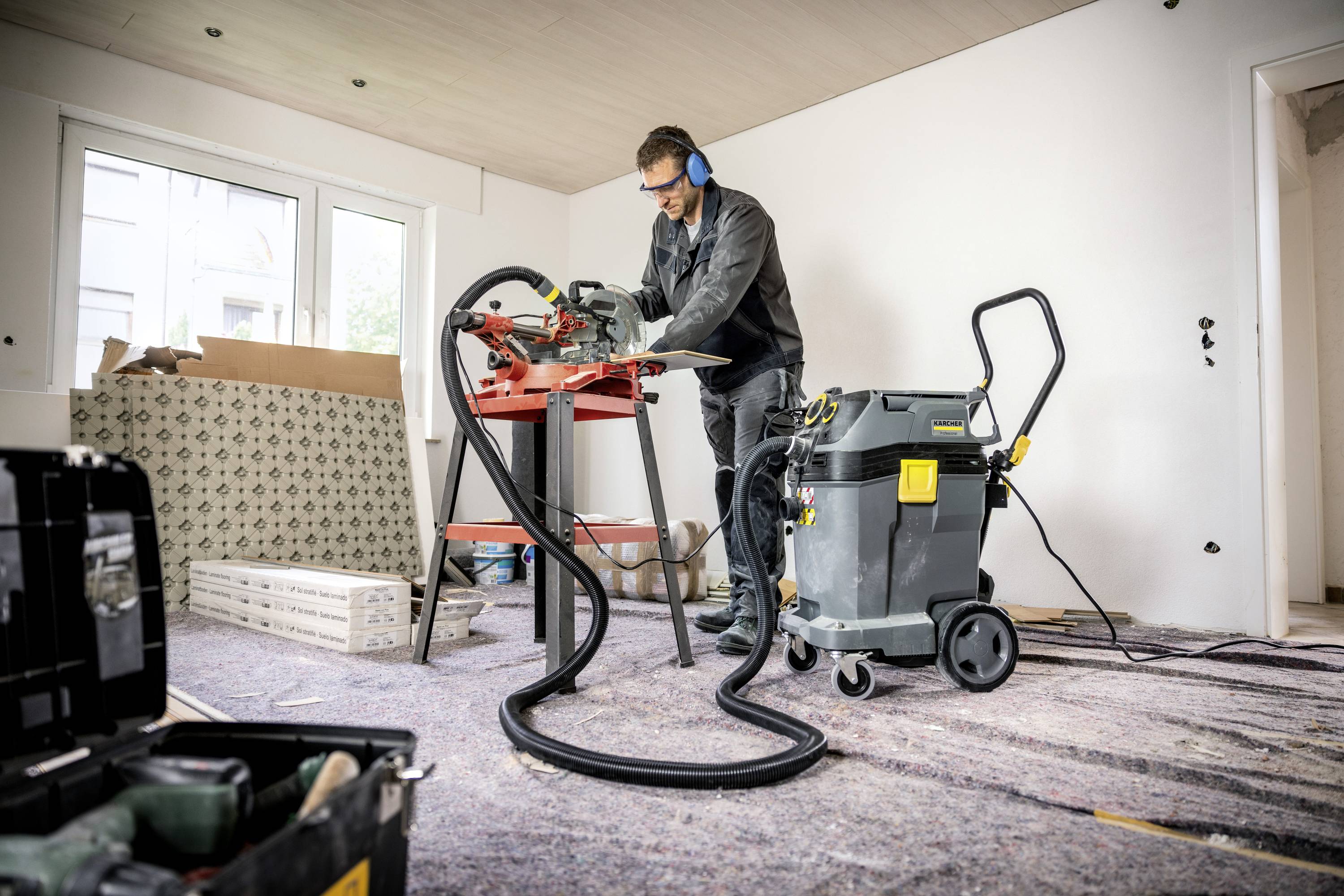 Mężczyzna pracuje w niedokończonym pomieszczeniu, używając piły na stole. Nosi odzież ochronną, a otoczenie przypomina plac budowy.