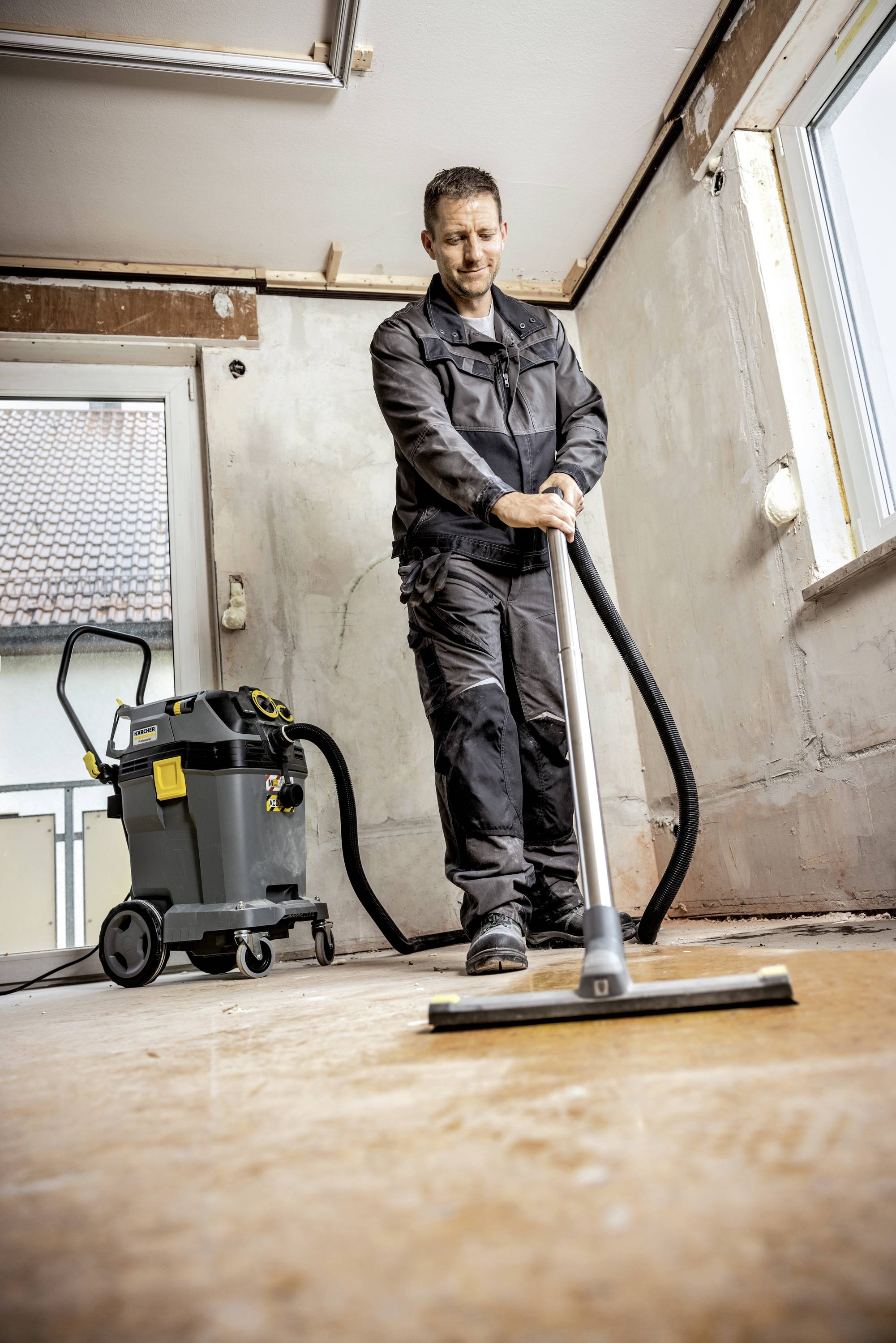 Osoba w ubraniu roboczym czyści podłogę pustego pomieszczenia przemysłowym odkurzaczem. Pomieszczenie wygląda na wymagające renowacji.