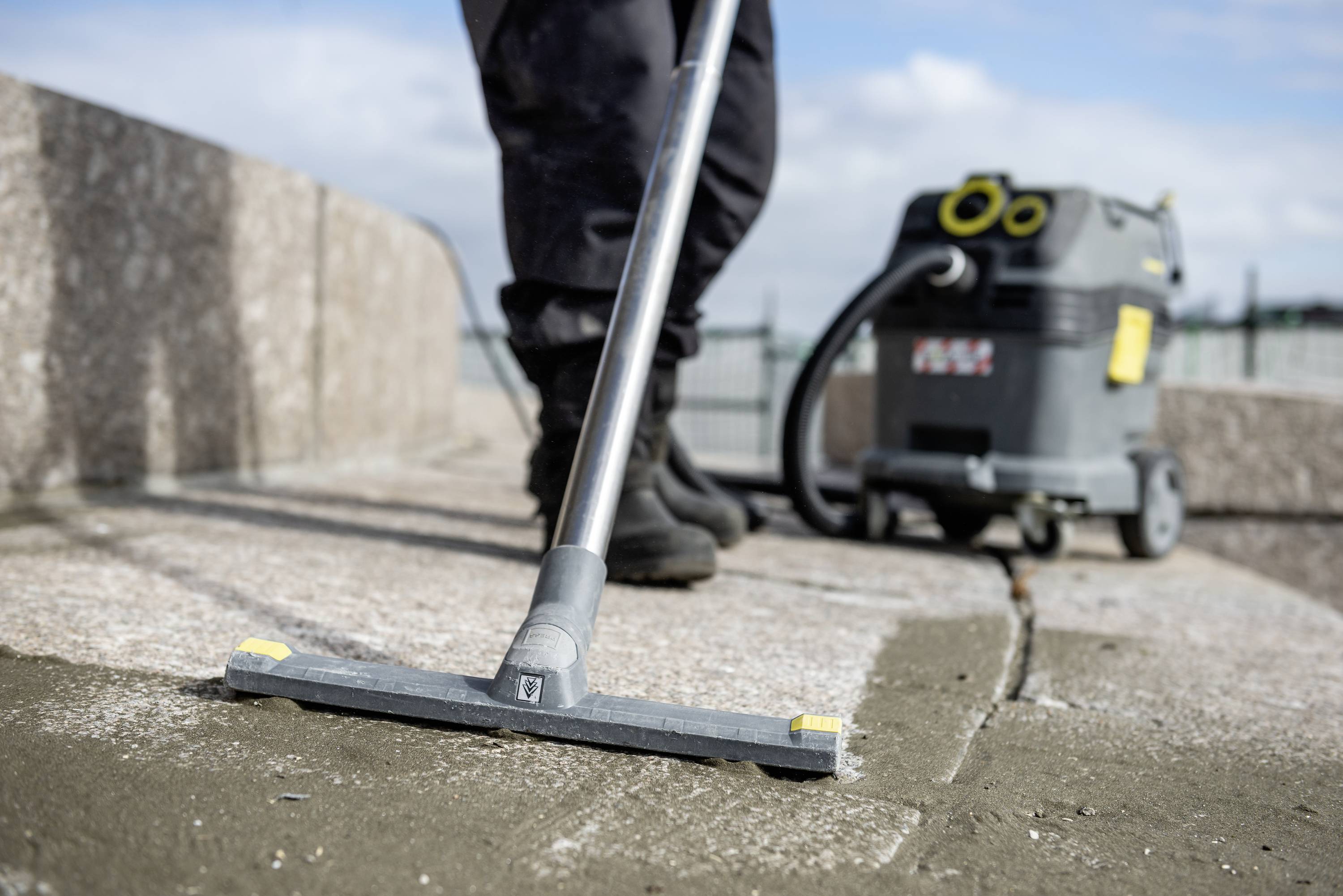 Osoba czyści kamienną powierzchnię przemysłowym odkurzaczem na wolnym powietrzu. W tle widoczne są błękitne niebo i chmury.