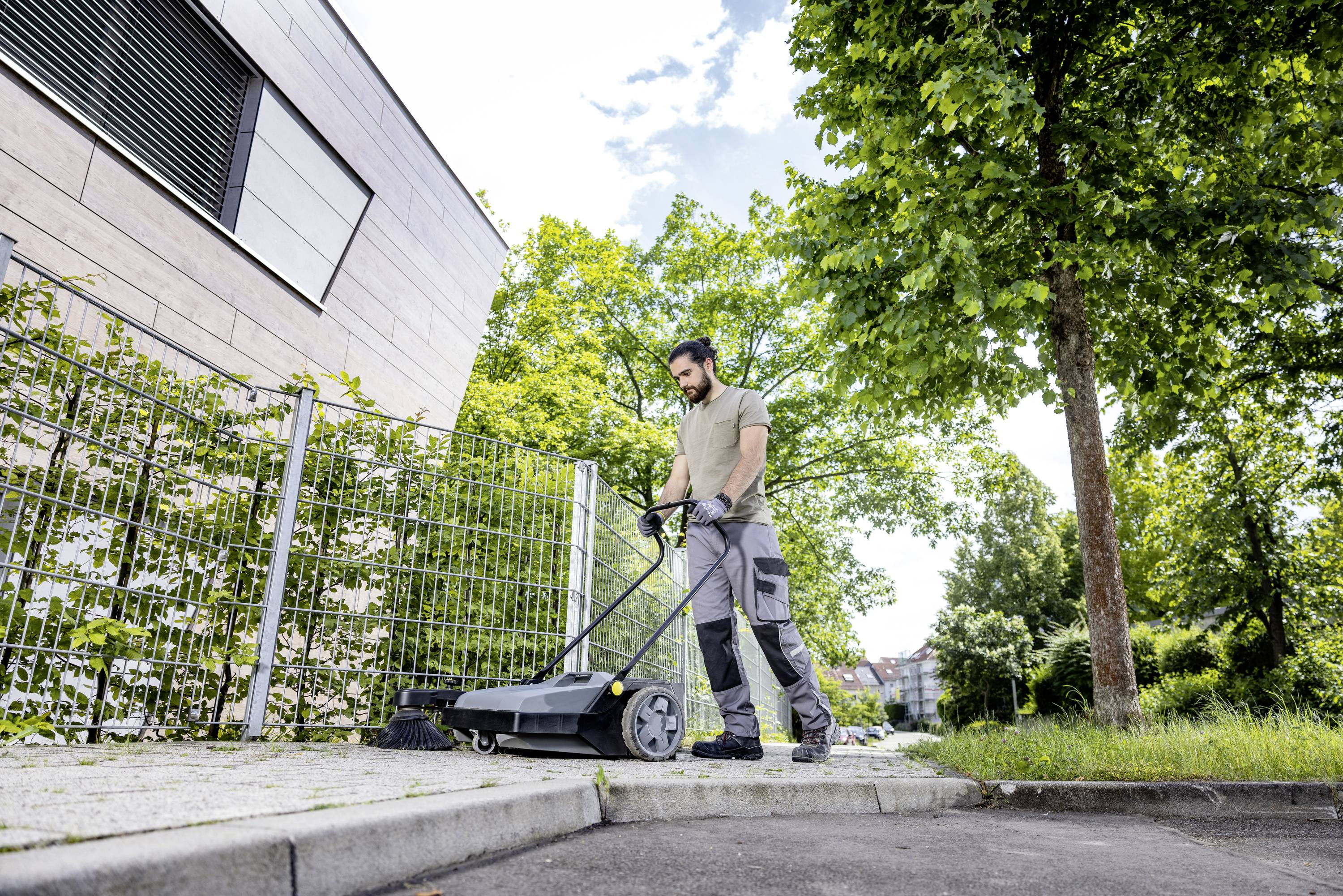 Mężczyzna jedzie zamiatarką wzdłuż chodnika przy nowoczesnym budynku. Drzewa obramowują krawędź chodnika.